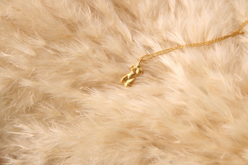Peut inclure: Un collier en or avec un petit pendentif en forme de silhouette humaine abstraite. Le collier est pos&eacute; sur un lit de plumes douces et blanches.