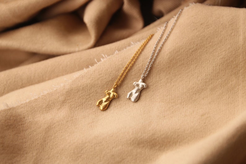 Peut inclure: Deux colliers en or et en argent avec un pendentif en forme de corps. Les colliers sont sur un fond en tissu brun.