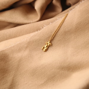 Peut inclure: Un collier en cha&icirc;ne dor&eacute;e avec un pendentif en or en forme de silhouette humaine. Le collier est pos&eacute; sur une surface en tissu beige.