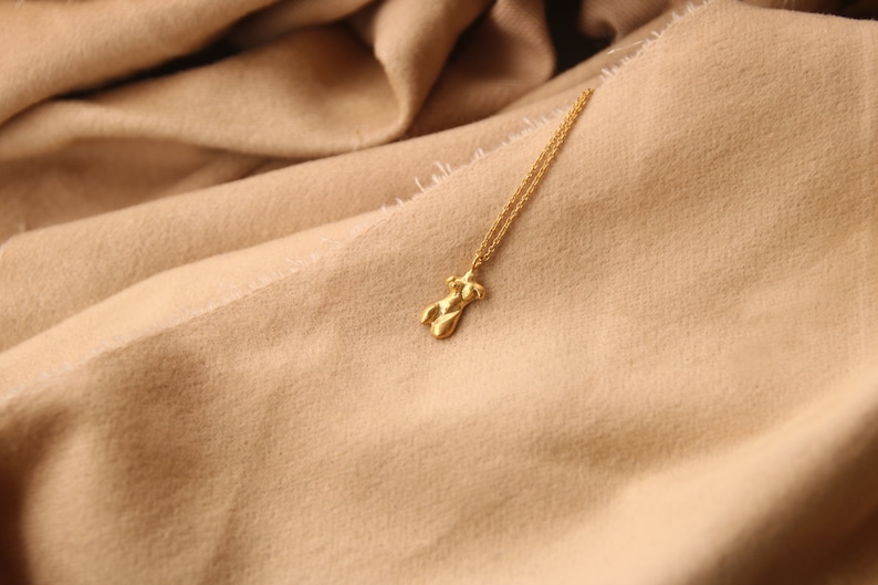 Peut inclure: Un collier en cha&icirc;ne dor&eacute;e avec un pendentif en or en forme de silhouette humaine. Le collier est pos&eacute; sur une surface en tissu beige.
