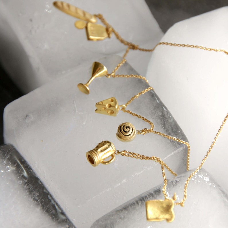 May include: Gold-colored charm necklaces featuring food and drink-themed pendants. The charms include a baguette, a wine glass, a cheese wedge, a cinnamon roll, a beer mug, and a rectangular shape. The necklaces are displayed on a textured, white surface.