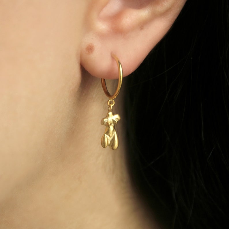 Puede incluir: Un pendiente de aro dorado con un peque&ntilde;o colgante con forma de torso femenino estilizado. El pendiente se muestra en primer plano, destacando los detalles de la joya. El dise&ntilde;o es simple y elegante, adecuado para el uso diario.