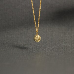 Op de afbeelding: Een goudkleurige ketting met een kleine, ronde hanger met een spiraalvormig ontwerp. De hanger hangt aan een fijne ketting. De achtergrond is effen donkergrijs.