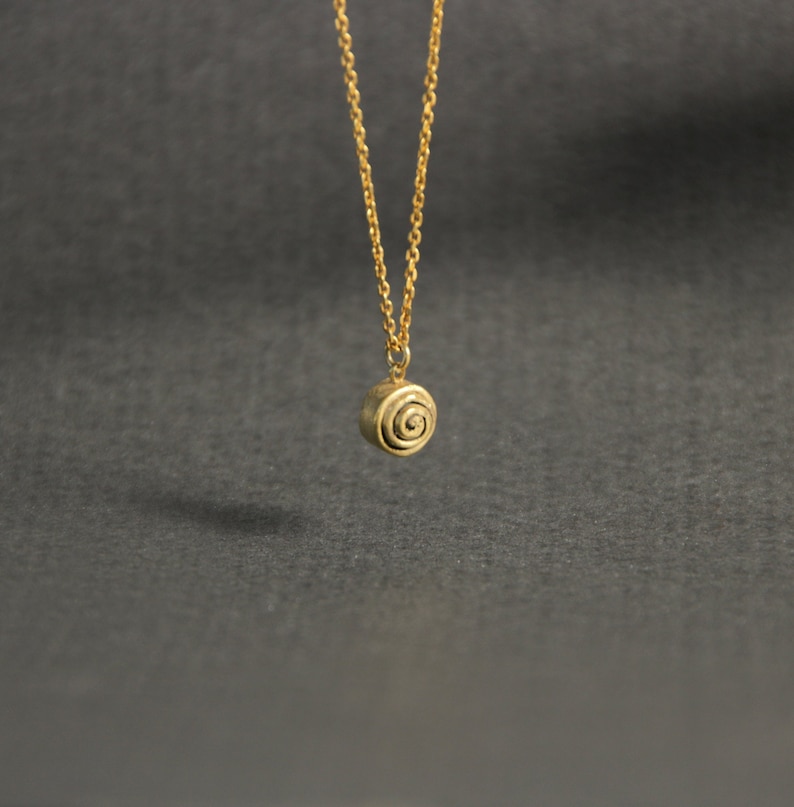 Op de afbeelding: Een goudkleurige ketting met een kleine, ronde hanger met een spiraalvormig ontwerp. De hanger hangt aan een fijne ketting. De achtergrond is effen donkergrijs.