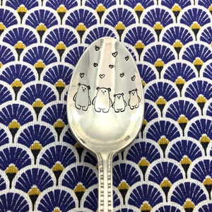 Peut inclure: Une cuillère en argent gravée de quatre ours et de cœurs. La cuillère est posée sur un tissu à motifs bleus et dorés.