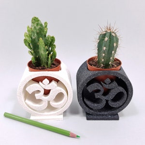 Pode incluir: Dois pequenos cactos em vasos em vasos brancos e pretos em forma de símbolo Om. Os cactos estão em vasos marrons.