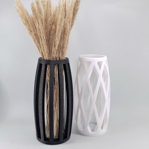 May include: Two decorative vases. One is black with vertical bars, holding dried wheat stalks. The other is white with a diamond lattice design. Both vases have a cylindrical shape and are on a white surface.