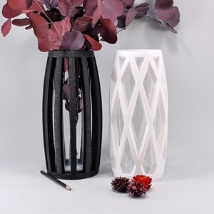 May include: Two decorative vases, one black and one white, with geometric designs. The black vase has vertical bars, while the white vase features a diamond pattern. Both vases are filled with burgundy-colored foliage. A pencil rests near the black vase.