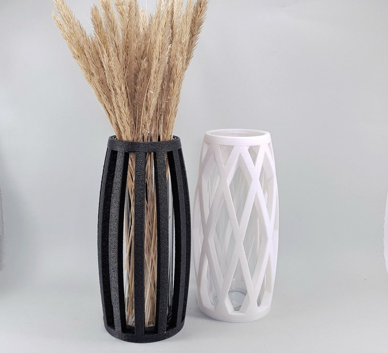 May include: Two decorative vases. One is black with vertical bars, holding dried wheat stalks. The other is white with a diamond lattice design. Both vases have a cylindrical shape and are on a white surface.