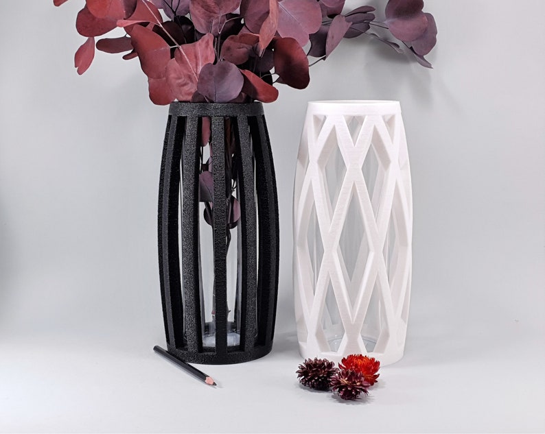 May include: Two decorative vases, one black and one white, with geometric designs. The black vase has vertical bars, while the white vase features a diamond pattern. Both vases are filled with burgundy-colored foliage. A pencil rests near the black vase.