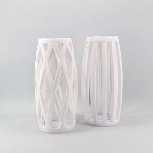 May include: Two white decorative vases. One vase has a geometric diamond pattern, and the other has vertical lines. Both vases have a clear glass interior, and are cylindrical in shape.