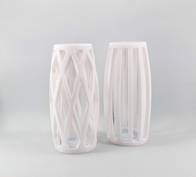 May include: Two white decorative vases. One vase has a geometric diamond pattern, and the other has vertical lines. Both vases have a clear glass interior, and are cylindrical in shape.