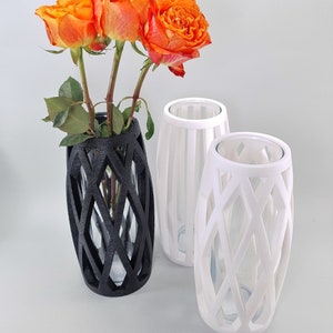 May include: Three decorative vases, two white and one black, with a geometric lattice design. The black vase holds orange roses. The vases have a clear glass insert. The vases are a modern home decor item.