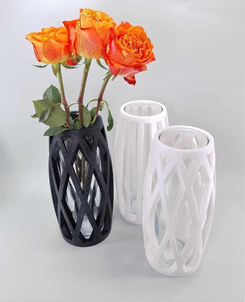 May include: Three decorative vases, two white and one black, with a geometric lattice design. The black vase holds orange roses. The vases have a clear glass insert. The vases are a modern home decor item.