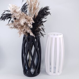 May include: Two decorative vases. One is black with a geometric lattice design, holding dried pampas grass in shades of beige, black, and brown. The other is white with vertical slats. Both vases are empty inside.