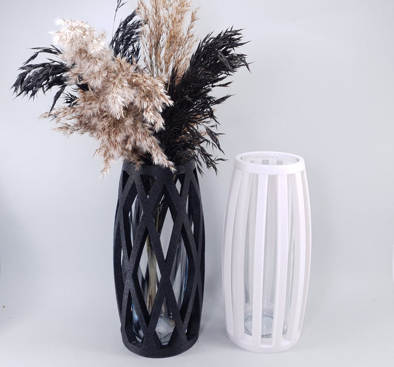 May include: Two decorative vases. One is black with a geometric lattice design, holding dried pampas grass in shades of beige, black, and brown. The other is white with vertical slats. Both vases are empty inside.