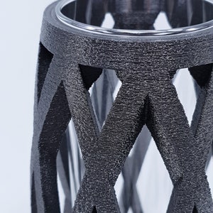 May include: A black, textured vase with a geometric lattice design. The vase has a clear glass insert, visible through the openwork. The top rim is also clear. The overall aesthetic is modern and decorative.