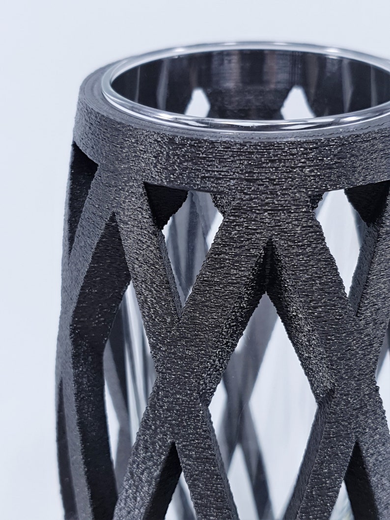 May include: A black, textured vase with a geometric lattice design. The vase has a clear glass insert, visible through the openwork. The top rim is also clear. The overall aesthetic is modern and decorative.