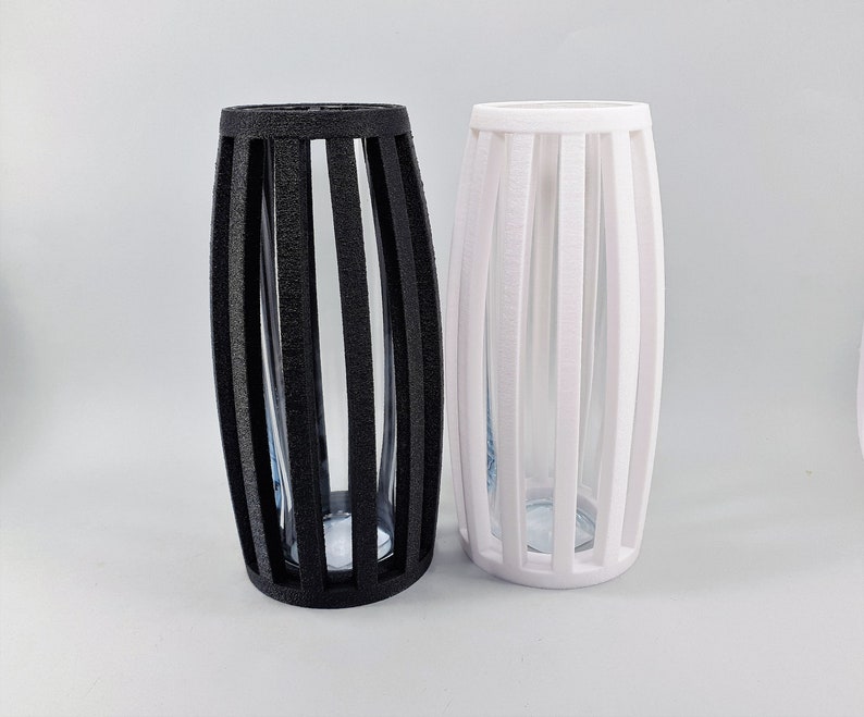 May include: Two decorative vases, one black and one white, with a vertical slat design. Each vase has a clear glass insert. The vases are cylindrical with an oval shape.