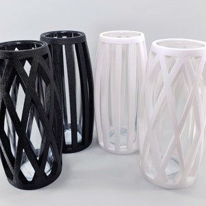 May include: Four decorative vases with clear glass inserts. Two are black with a geometric lattice design, and two are white with a vertical slat design. The vases have a cylindrical shape with a slightly wider base.