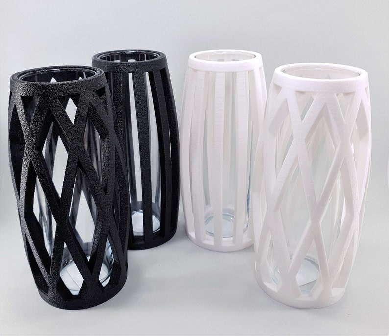 May include: Four decorative vases with clear glass inserts. Two are black with a geometric lattice design, and two are white with a vertical slat design. The vases have a cylindrical shape with a slightly wider base.