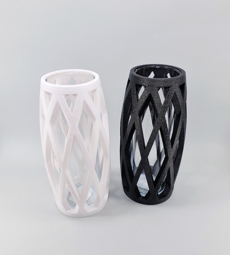 May include: Two decorative vases. One is white, the other is black. Both have a lattice design around a clear glass interior. The vases are cylindrical with an oval shape.