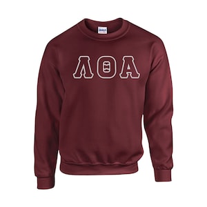 May include: A maroon crewneck sweatshirt with the letters ΛΘA in white, outlined in maroon. The letters are centered on the chest. The sweatshirt is made of a soft material and has a ribbed collar, cuffs, and hem.