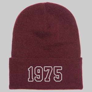 May include: A maroon beanie with the year "1975" embroidered in white block letters. The hat is made of a knitted material and has a folded brim. The beanie is a classic style, suitable for cold weather.