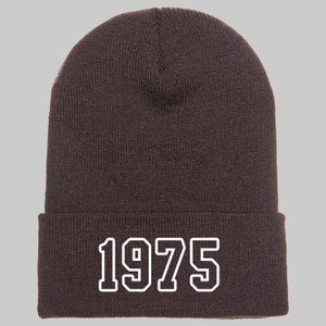 May include: A dark brown beanie with the year "1975" in white block letters across the front. The beanie is made of a knitted material and has a folded brim. The hat is a casual accessory.