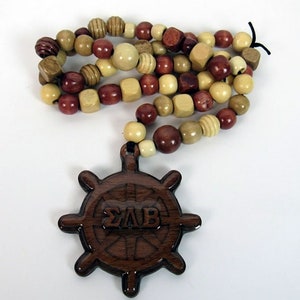 May include: A wooden beaded necklace with a dark brown ship's wheel pendant. The beads are various shapes and colors, including light tan, dark red, and brown. The pendant has the letters ΣΛΒ carved into it.