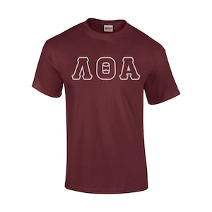 May include: Maroon short-sleeve t-shirt with the letters ΛΘA in white, outlined in maroon. The shirt is made of a soft material and has a crew neck. The brand name "Gildan" is visible on the tag.
