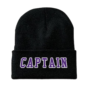 May include: Black knit beanie with the word "CAPTAIN" in purple block letters with a white outline. The beanie is a classic style, suitable for cold weather. The text is centered on the folded brim.