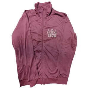 May include: A maroon zip-up track jacket with a high collar and ribbed cuffs. The jacket features white embroidered text on the chest: "ΛΘA 1975". The jacket is made of a smooth, shiny material.