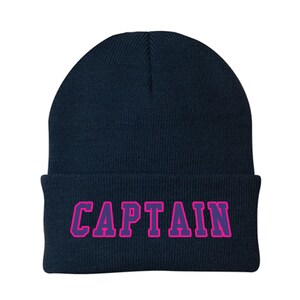 May include: A navy blue knit beanie with the word "CAPTAIN" in purple letters outlined in pink. The beanie is a classic style, suitable for cold weather.