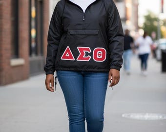 May include: A black quarter-zip hooded windbreaker featuring the Greek letters Delta Sigma Theta in red with white outlines on the front chest, paired with blue denim jeans and white sneakers. The jacket has a front pocket and drawstring hood.