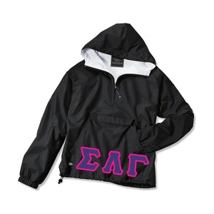 May include: Black hooded anorak jacket with a white interior lining. The jacket features a front pocket and a drawstring at the hem. The letters ΣΑΓ in purple with a pink outline are on the front. The jacket is made by Charles River.
