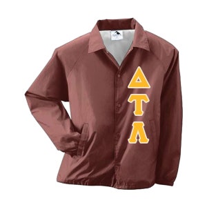 May include: A maroon coach jacket with a button-up front, collared neckline, and two front pockets. The jacket features the Greek letters Delta, Tau, and Lambda in a gold color on the front.