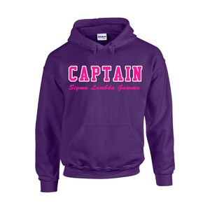 May include: A purple hooded sweatshirt with the word "CAPTAIN" in white block letters outlined in pink. Below the word "CAPTAIN" is the text "Sigma Lambda Gamma" in pink script. The sweatshirt has a front pocket and a hood.