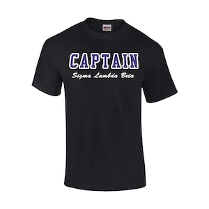 May include: Black t-shirt with the text "CAPTAIN" in white with a purple outline and the text "Sigma Lambda Beta" in white below.