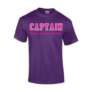 May include: A purple t-shirt with the word "CAPTAIN" in white outlined pink block letters across the chest. Below the word "CAPTAIN" is the text "Sigma Lambda Gamma" in pink script. The shirt is made of a soft material.