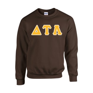 May include: A brown sweatshirt with the Greek letters Delta, Tau, and Lambda in gold with a white outline. The letters are centered on the chest of the long-sleeved crew neck top.