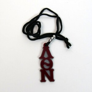 May include: A maroon pendant necklace with the Greek letters A, Θ, and N. The pendant hangs from a black cord with silver beads. The necklace is displayed on a white background.