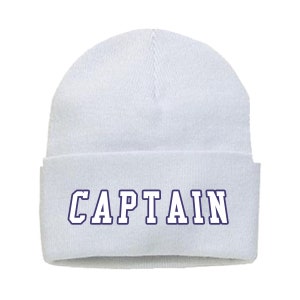 May include: A white beanie with a folded brim. The word "CAPTAIN" is printed across the front in navy blue block letters with a white outline. The hat is made of a soft, knitted material.