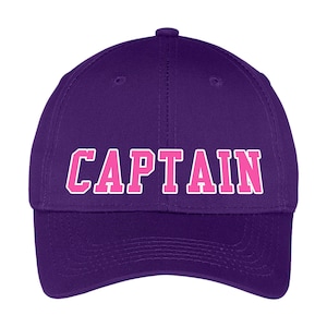 May include: A purple baseball cap with the word "CAPTAIN" in pink block letters outlined in white. The hat has a curved brim and a button on top. This hat is a great accessory for anyone.