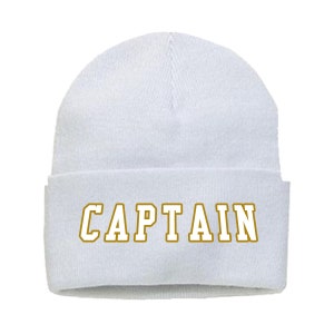 May include: A white beanie with the word "CAPTAIN" in gold lettering across the front. The beanie is a classic, folded-brim style, suitable for cold weather. The text is in a bold, blocky font.