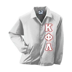 May include: A white coach jacket with a button-up front, collared neckline, and side pockets. The jacket features the Greek letters K, Φ, and Λ in red on the front. The jacket is made of a lightweight material.