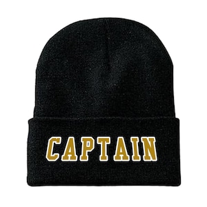 May include: Black beanie hat with the word "CAPTAIN" in gold block letters with a white outline. The hat is a classic, knitted style, suitable for cold weather. The text is centered on the folded brim.
