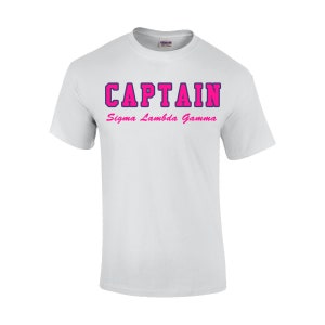 May include: White t-shirt with the word "CAPTAIN" in a bold, pink font with a dark blue outline. Below the word "CAPTAIN" is the text "Sigma Lambda Gamma" in a smaller, pink font. The shirt is made of a soft material.