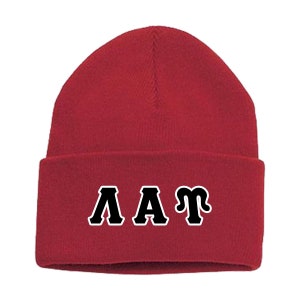 May include: A red beanie with the letters ΛAT in black with a white outline. The hat is made of a soft, knitted material and has a folded brim. The beanie is a warm accessory for cold weather.
