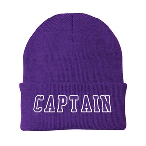 May include: A purple beanie hat with the word "CAPTAIN" in white block letters across the front. The hat is made of a soft, knitted material and has a folded cuff.